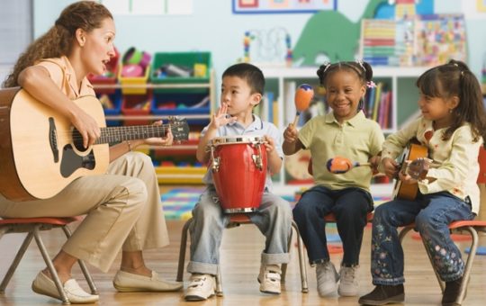Adult playing guitar and three young children playing a bongo, maracas and guitar