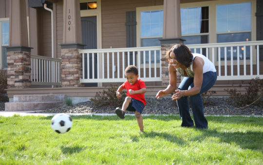 boys kicks soccer ball with woman watching and smiling