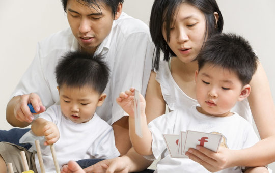 Two boys sit in their mother and fathers' laps and play cards.