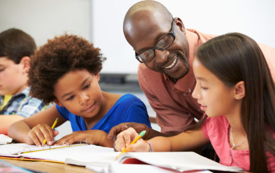 A man leans over two pupils' shoulders and smiles encouragingly as they show him their work