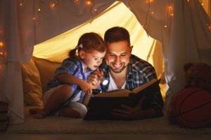 a father and daughter sit under an indoor tent, reading a book and smiling