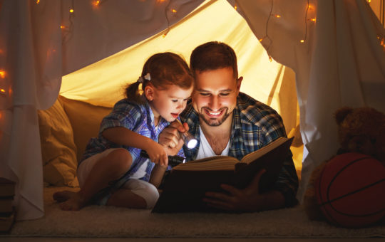 a father and daughter sit under an indoor tent, reading a book and smiling