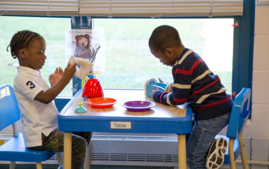 Two young boys play with dishes and other kitchen items at a table in their classroom.