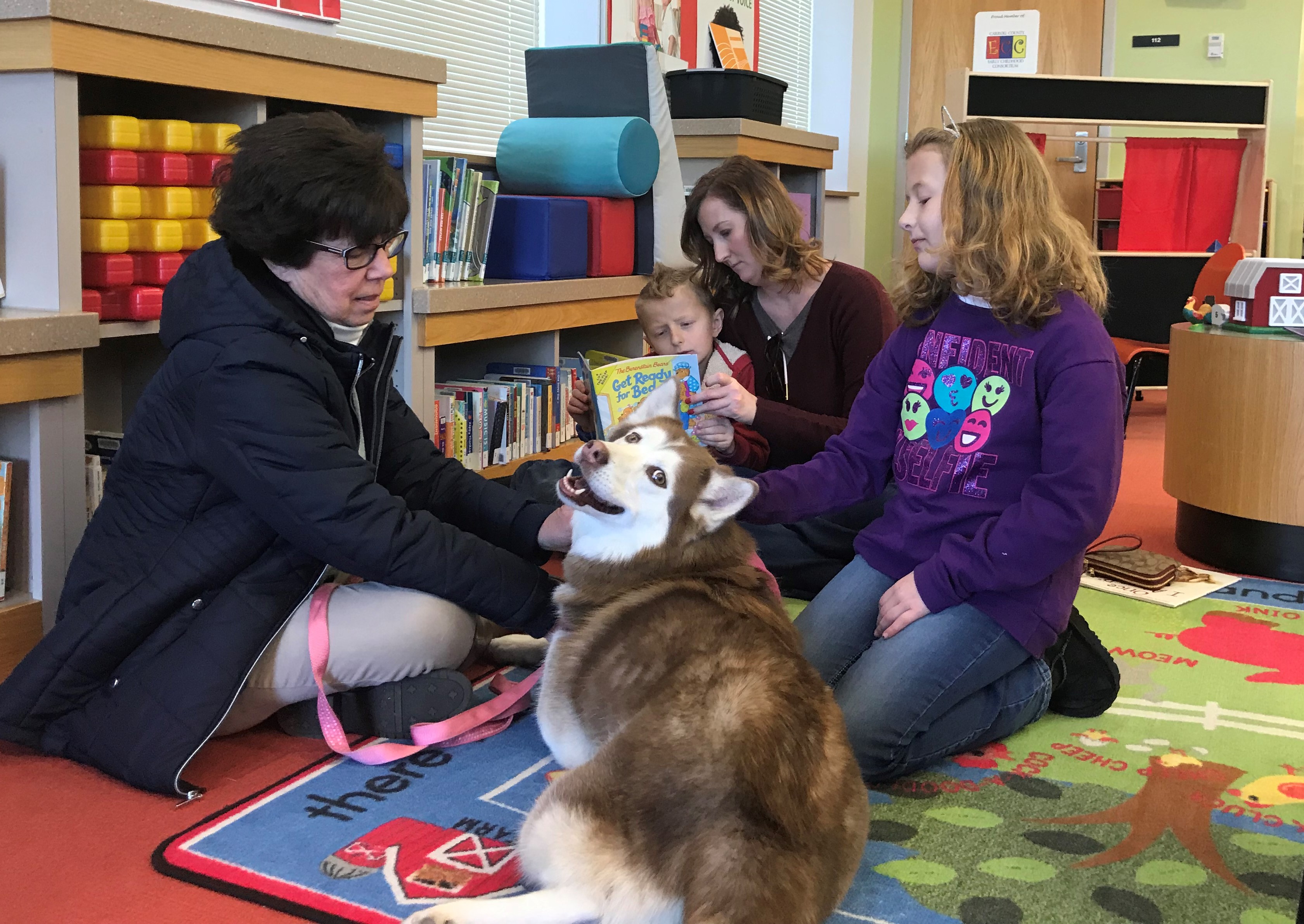 A boy reads to a dog at the library while his mom and sister listen