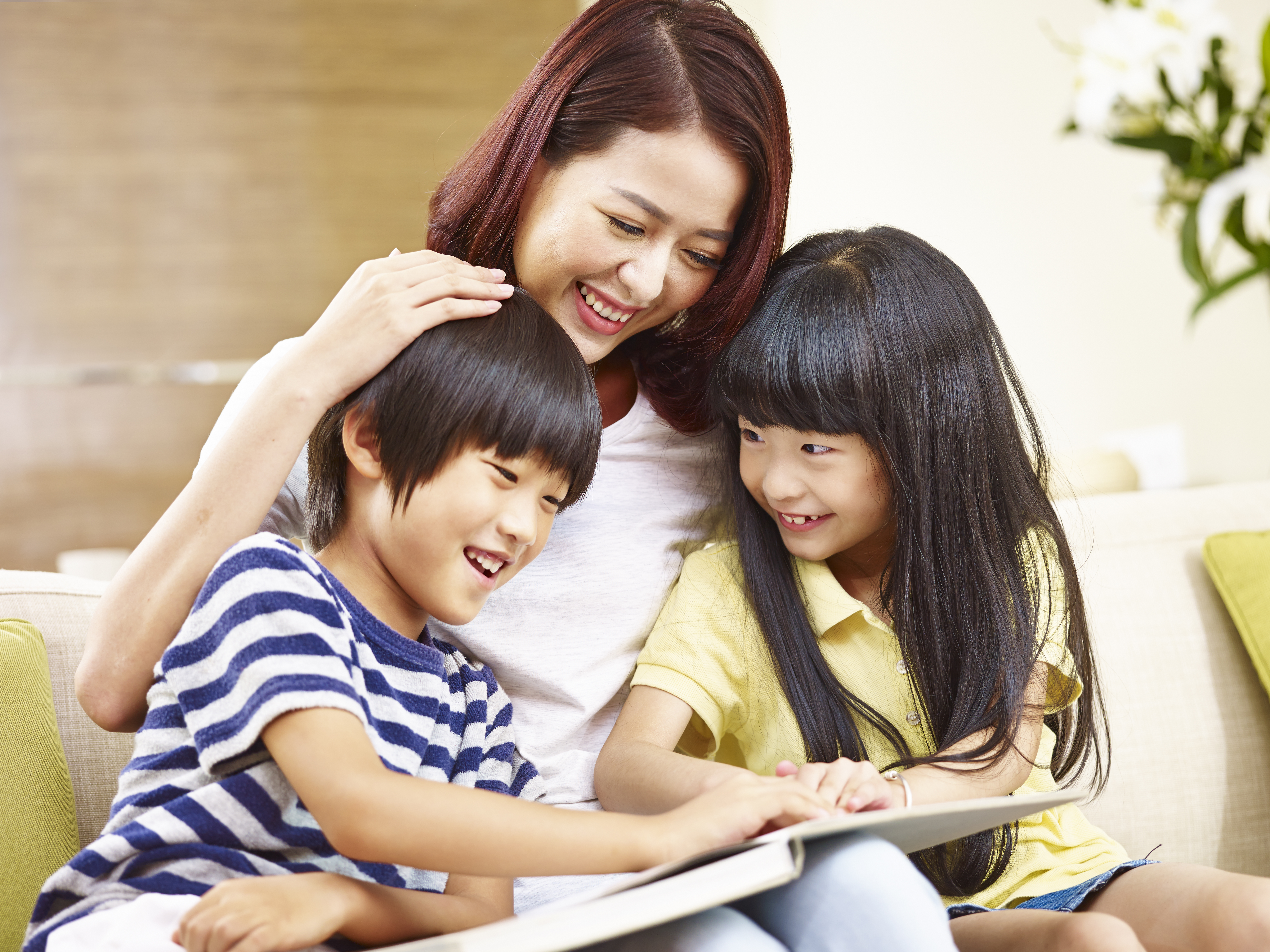 young asian mother sitting on couch at home reading story to two little children.