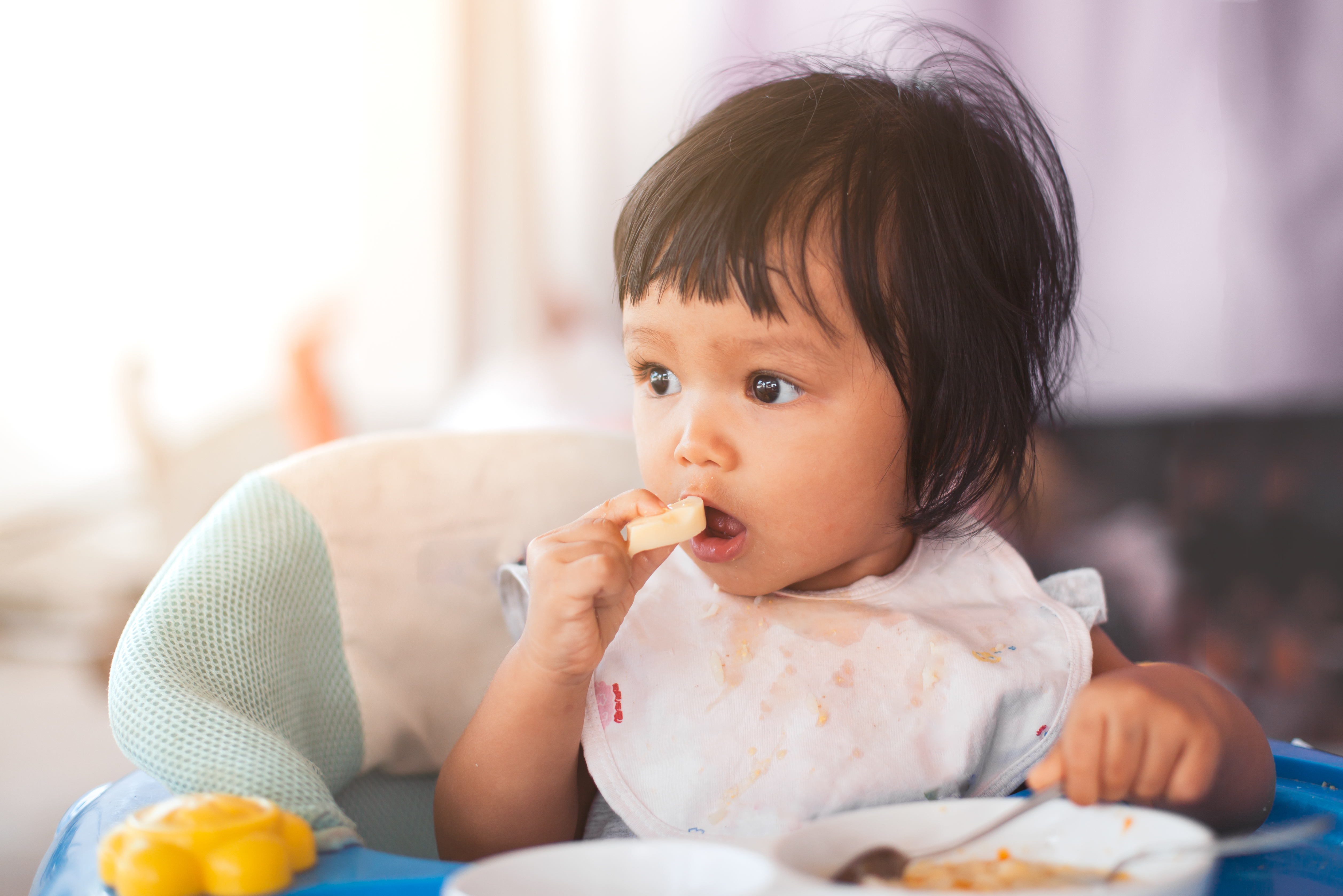 Cute baby asian child girl eating healthy food by herself and making a mess on her face and hand