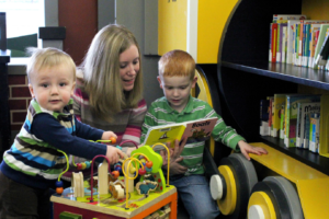 woman reading to young boy while toddler plays with activity table at library