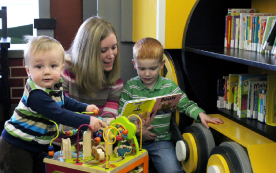 woman reading to young boy while toddler plays with activity table at library