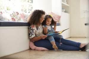 A mother sits on a cushion the floor, reading to her young daughter