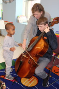 a young man plays the cello to a young boy while teacher assists