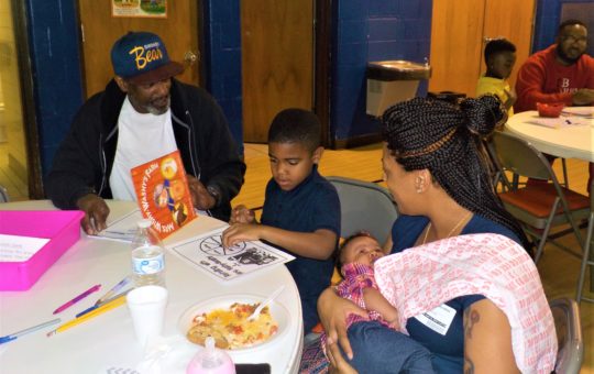 An African American man sits at a table with the book Mrs. Wishy Washy with his son and son's mother who is holding an infant girl