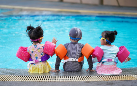The backs of three young children in goggles and arm floats sitting on the edge of a pool