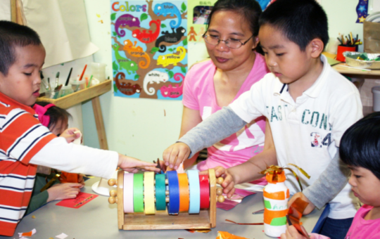 teacher sits at table with three children doing crafts with colorful tape