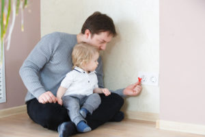 father sits on the floor with a toddler on his lap while putting in safety caps on the electrical socket