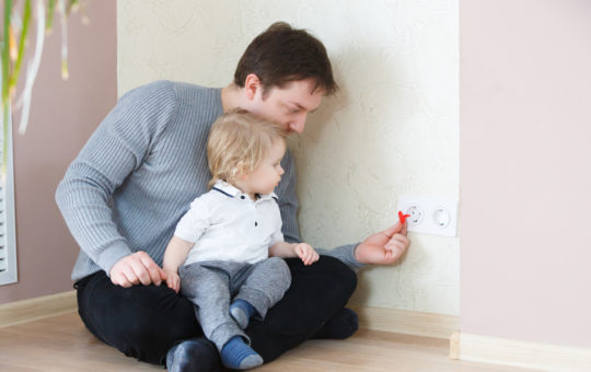 father sits on the floor with a toddler on his lap while putting in safety caps on the electrical socket