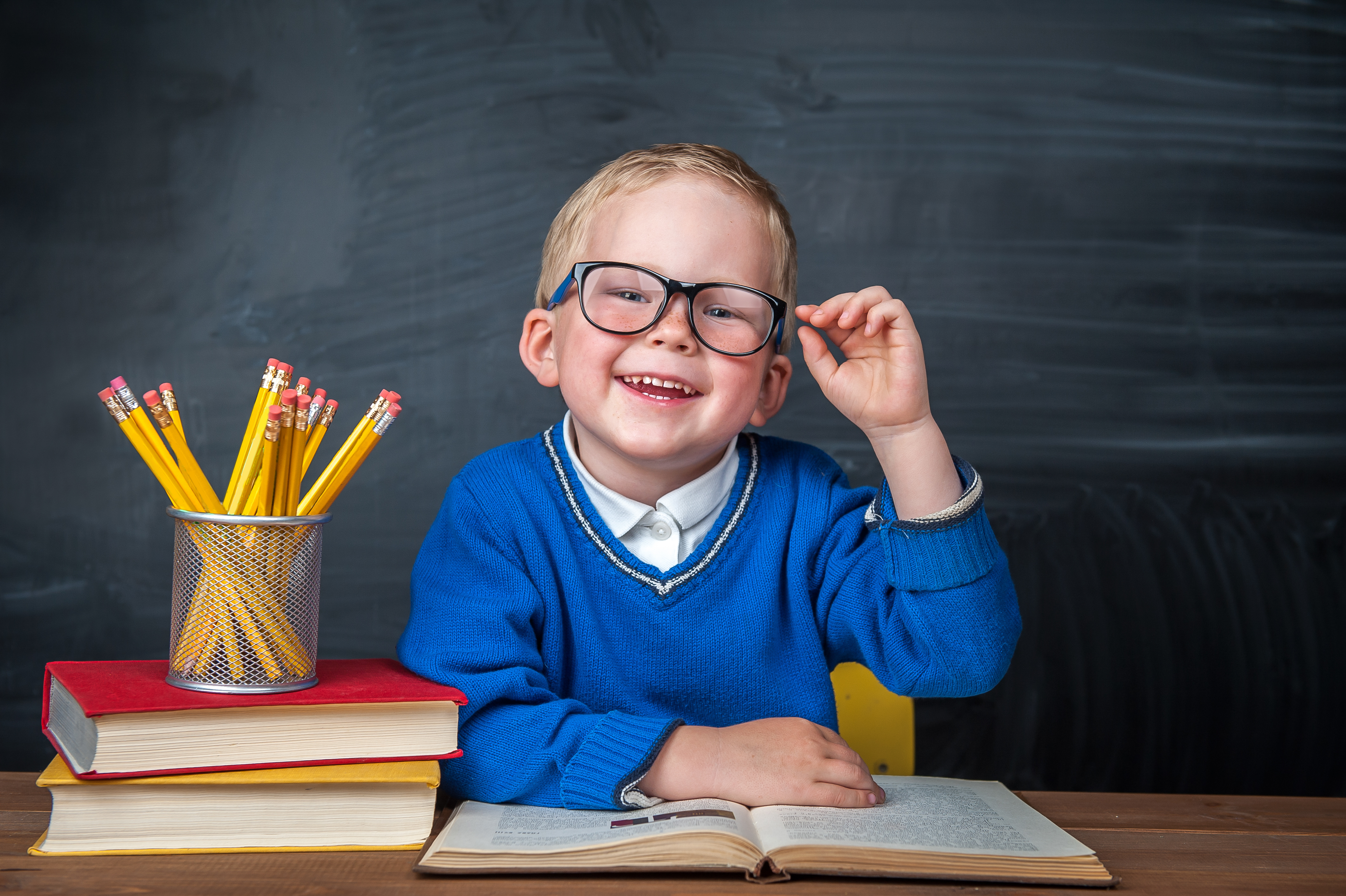 boy touching his glasses sitting at a desk with large books and container of pencils