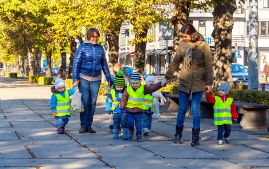 Two adults are walking hand in hand with five children wearing safety vests on the sidewalk
