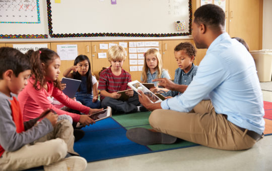 An african american teacher sits on the floor using a tablet with a group of children who are also holding tablets