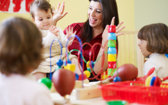 Female teacher smiles with hands in the air with a toddler girl sitting on her lap as two other children sit with table toys