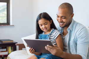 African American father and smiling daughter sitting on sofa using digital tablet. Happy dad watching cartoon with his cute little girl. Cheerful father and daughter sitting on couch at home and playing with computer.