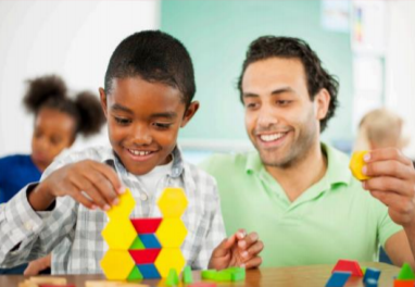male teacher sits beside school age african american boy while stacking shapes