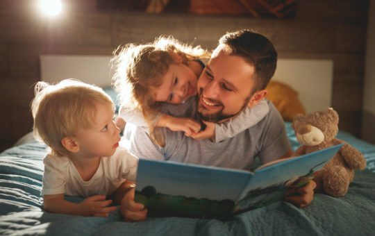 father, daughter and son lie on the bed reading a book together