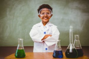 Boy wearing lab coat stands with arms crossed in front of a table with beakers filled with colored liquid