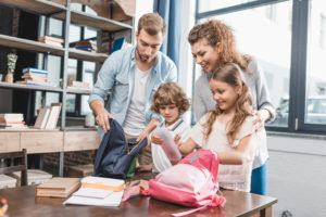 mother and father watch over the shoulders of son and daughter as they pack their backpacks