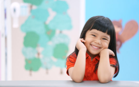 Asian girl smiles while sitting at table with her face resting on her hands